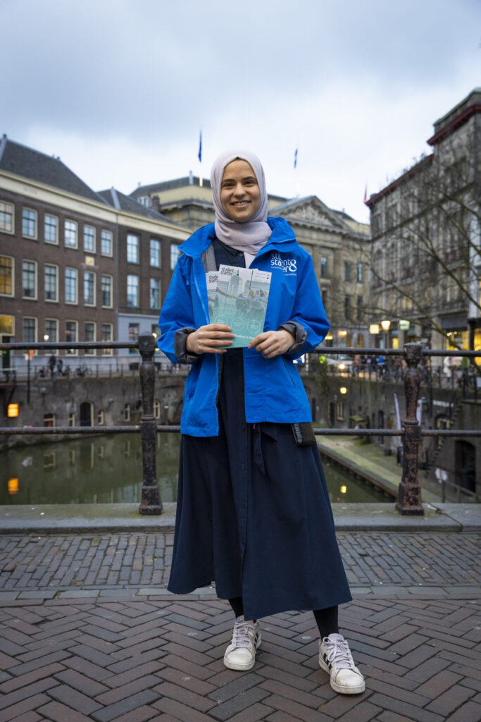 Esma staat met flyers in haar hand en een blauwe jas aan voor het stadhuis