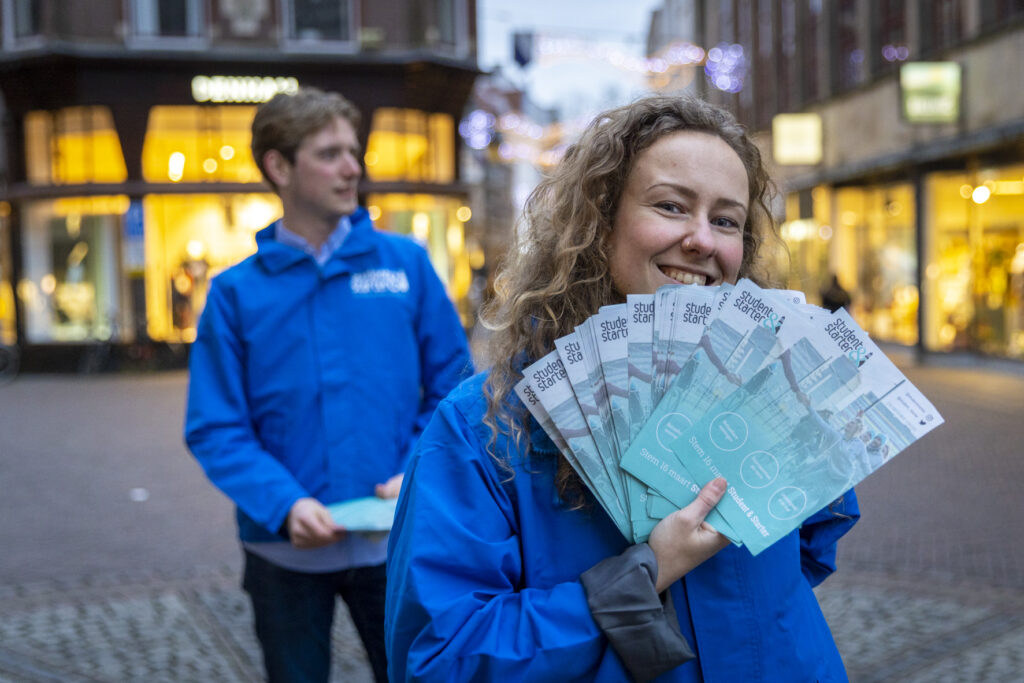 Remi presenteert lachend flyers met Rob op de achtergrond