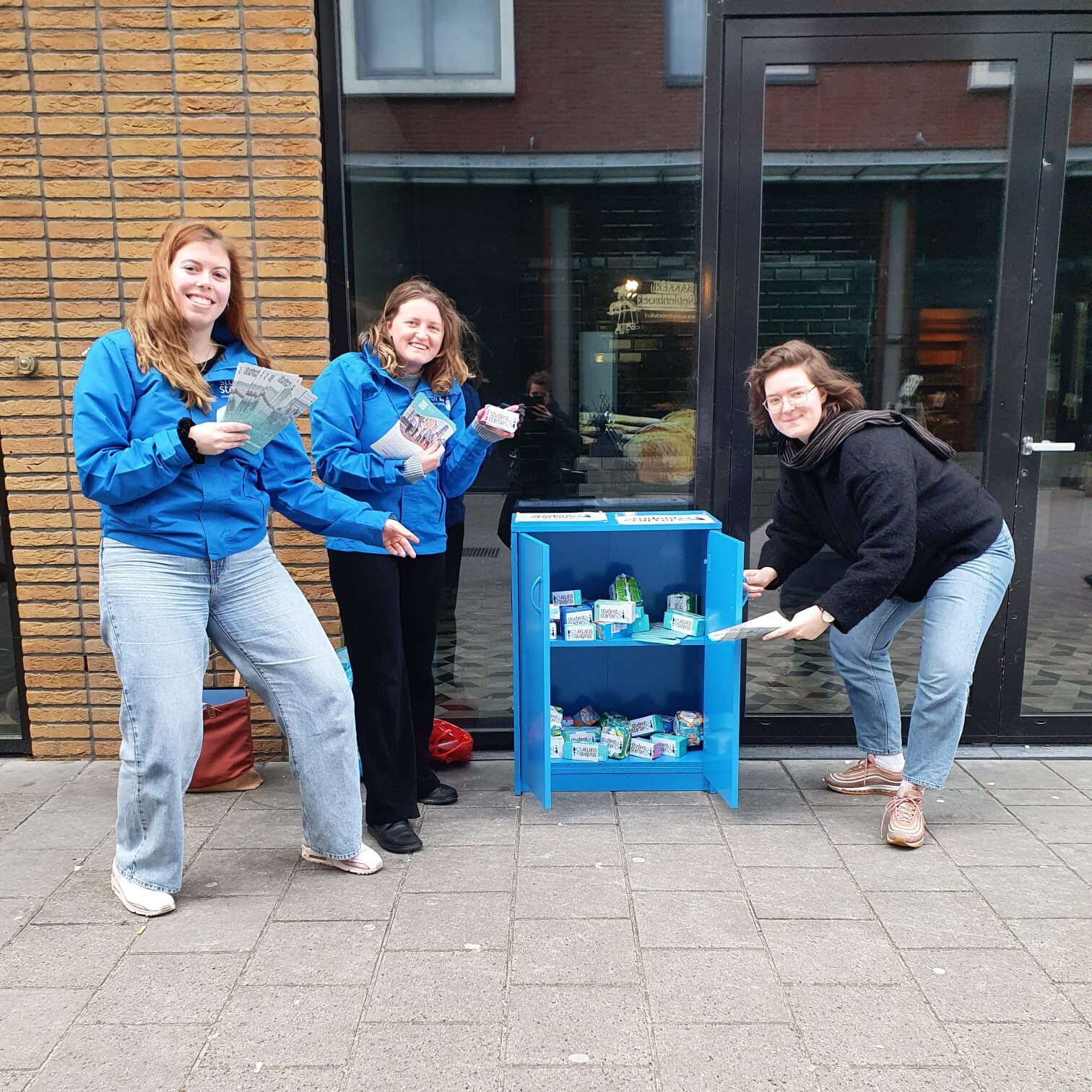 Myrthe, Boukje en Sabrina staan lachend voor een kast met gratis menstruatieproducten